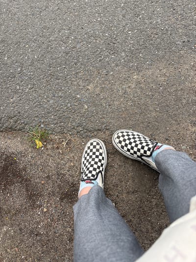 This is an aerial shot of a person's feet wearing black and white checkered slip-on Vans sneakers and gray pants standing on an asphalt surface in Větrušice, Czechia. The asphalt has a rough texture with visible small stones and pebbles. A small patch of green grass with some dry twigs is visible to the left of the left shoe. The lighting suggests it is daytime.