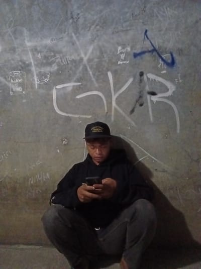 A young man is crouched down in front of a concrete wall covered in graffiti. He is wearing a black hoodie and dark pants, and a black baseball cap. He is looking at his phone, which he holds in his hands. The graffiti on the wall is a mix of spray paint and chalk, and includes words, symbols, and abstract shapes. Some of the graffiti appears to be in Indonesian. The lighting is dim, casting shadows on the wall and the man. The image appears to have been taken in the city of Bojong Gede, Indonesia.