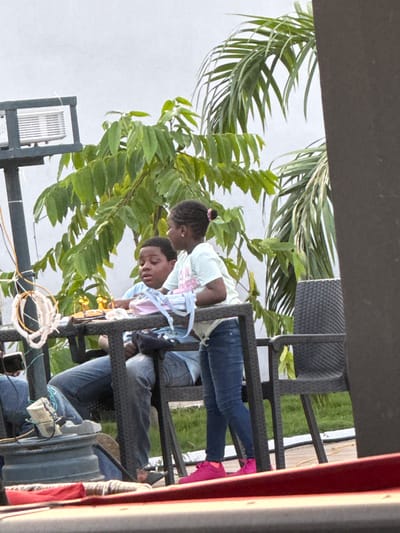 In Rubuchi, Nigeria, two children are seated at a black rattan-style table outdoors, with lush green foliage and palm fronds framing the background. A young boy, wearing a light blue shirt, sits on the left, looking intently at something on the table. Beside him, a young girl in a white t-shirt and blue jeans stands, holding a light blue object, possibly fabric or paper, and appears to be interacting with the boy. On the table, a small orange and gold object with two points, resembling a small trophy or decoration, is visible, along with a pink and light blue bag. The children are seated on dark chairs, and the scene is illuminated by daylight. In the foreground, a red surface suggests another seating area or platform. The presence of a projector-like device mounted on a pole in the background hints at an entertainment setup.