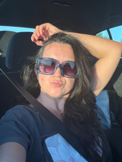 The image shows a woman lying down in a car, looking at the camera. She has long, dark hair and is wearing large, square sunglasses with purple lenses. Her mouth is slightly open, and she appears to be smirking or pouting. The sunlight is casting shadows on her face, suggesting it is daytime. The background shows the interior of a car, with a black seat and headrest. Through the window, trees and a blue sky are visible, indicating an outdoor setting. A grey seatbelt is stretched across her chest. The overall mood seems relaxed and casual, possibly taken during a road trip or while parked. The visible text on her shirt is a stylized floral design.