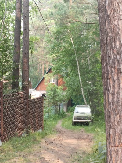 The image depicts a rural setting in the city of Bibikha, Russia. A light green vehicle, possibly a Lada Niva, is parked on a dirt path in front of a wooden house with a red roof. The vehicle is facing the camera. To the left of the path, there is a decorative metal fence. The environment is densely wooded with tall trees and lush green foliage. The lighting suggests it is daytime, with soft, diffused light filtering through the trees. The overall impression is one of a secluded, natural environment.