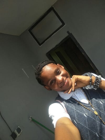 A young individual, appearing female, with dark skin and short dark braided hair, smiles directly into the camera while taking a selfie in Cotonou, Benin. They wear a white collared shirt beneath a dark gray or blue argyle-patterned V-neck sweater vest. Multiple dark beaded bracelets adorn their left wrist, and a long gold-toned chain with a medallion hangs around their neck. The subject's right hand is positioned with fingers resting under their chin. The background consists of a light gray painted wall or ceiling. Two dark, rectangular openings are visible: one higher and centered, and another vertical opening, likely a doorway, to the right. Exposed dark electrical wiring leading to a white wall fixture and a green horizontal pipe are visible on the lower left wall. The scene is brightly lit from the upper right, casting a shadow under the subject's chin.