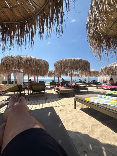 The image depicts a beach scene in Limenaria, Greece, under a clear blue sky. In the foreground, a person's legs are visible, lying on a dark lounge chair. The sandy beach is dotted with several thatched umbrellas providing shade over rows of lounge chairs. Some chairs are occupied by people, who are either sitting or lying down, and some have towels draped over them. In the background, the calm, blue sea stretches out to the horizon, with a few small boats visible. The overall atmosphere is relaxed and suggests a typical day at a beach resort.