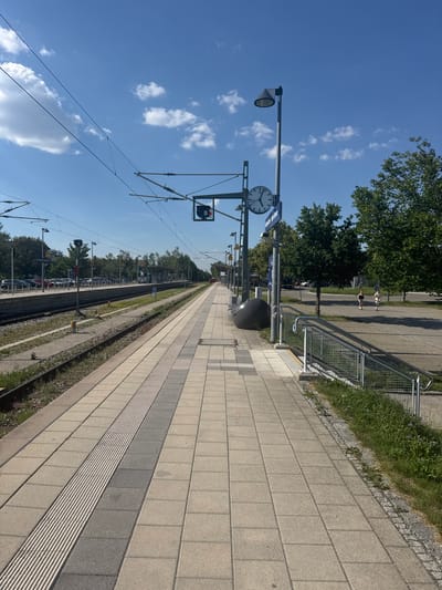 The image depicts a train station platform on a sunny day. The perspective is looking down the platform, with a clear blue sky and scattered white clouds overhead. The platform itself is paved with light-colored square tiles and features a tactile paving strip. To the left of the platform are train tracks, with overhead power lines stretching across the sky.

Several objects are visible on the platform, including a lamppost with a light fixture, a large circular clock with black hands and Roman numerals, and a dark, rounded trash receptacle. A metal railing with a wire mesh barrier runs along the edge of the platform, separating it from a grassy area with some small purple flowers. In the distance, trees and buildings are visible, suggesting an urban or suburban setting.

Two people are seen walking on the paved area beyond the railing, appearing as small figures in the distance. One person is wearing a light-colored outfit, and the other is in darker clothing. The overall scene conveys a sense of calm and ordinary day at a train station. The time appears to be sometime in the late morning or early afternoon, judging by the bright sunlight and the position of the shadows. No discernible text is clearly legible in the image, although some faint markings might be present on signs in the distance. The location is identified as Höhenkirchen-Siegertsbrunn, Germany.