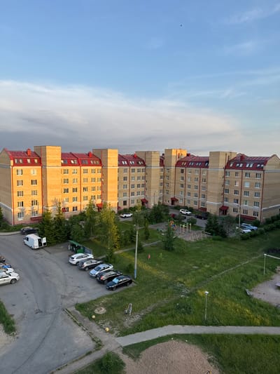 The image captures an aerial view of a residential area in Kirishi, Russia, likely taken during late afternoon or early evening, as indicated by the warm, golden light illuminating the buildings. The dominant feature is a long, multi-story apartment building constructed with light yellow bricks and a distinctive red-tiled, mansard-style roof. The building is segmented into several identical sections, each with multiple floors, numerous windows, and small dormer windows on the upper level.
In front of the building, a paved area serves as a parking lot where several cars are parked, including a white van with its rear doors open. Adjacent to the parking area is a strip of green grass, interspersed with trees and bushes. A children's playground with swings and colorful structures is visible within the green space. Further to the right, more grass and trees are present, along with streetlights and a pathway. The sky above is a clear blue with wispy white clouds, and a small bird is visible in flight. The overall scene depicts a typical urban residential environment with modern architecture and common amenities.