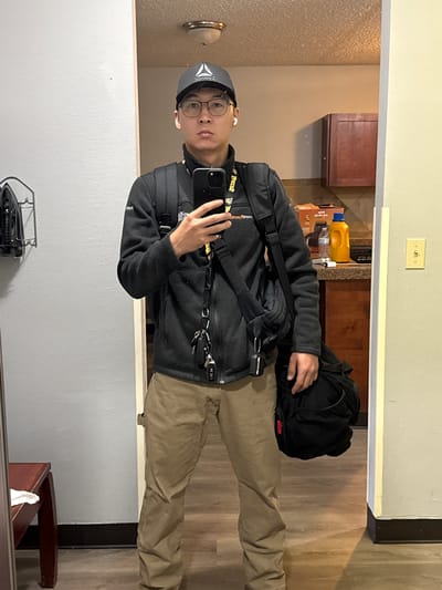 A full-body, front-facing mirror selfie shows a young adult male wearing a black Reebok baseball cap, clear-rimmed glasses, and a black zip-up fleece jacket over a yellow lanyard with some writing on it. He is holding a black smartphone in his right hand, taking the photo. He has a black backpack on his shoulders and a black messenger bag slung over his left shoulder. A set of keys hangs from the zipper of his jacket. He is wearing tan pants. The background shows a hallway with a mirror on the left wall, an ironing board and iron on the wall, and a doorway to a kitchen area on the right. The kitchen has wooden cabinets, a countertop with a yellow cleaning spray bottle, and a light fixture on the ceiling. The floor in the hallway appears to be wood laminate. The image is taken indoors, in what appears to be a residential setting. The location is not specified to be Vancouver, United States.