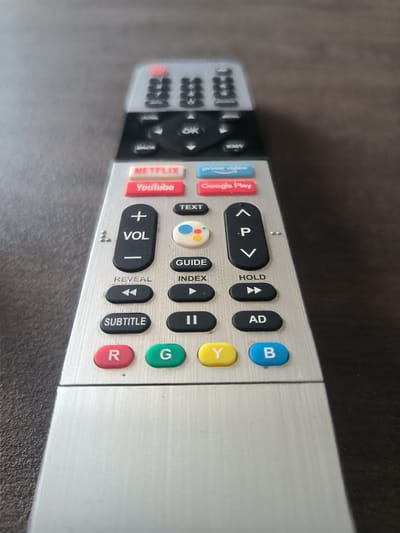 This is a close-up, eye-level shot of a silver television remote control resting on a dark brown, wooden surface. The remote is positioned vertically and slightly angled towards the viewer. The background is blurred, indicating a shallow depth of field, and the focus is sharply on the remote's buttons and labels.
The remote has a brushed silver finish with several black buttons arranged in a grid-like pattern. Prominent among these are dedicated app buttons for Netflix, YouTube, Prime Video, and Google Play, each with their respective logos or text. There are also standard navigation buttons, including up, down, left, and right arrows, an "OK" button, volume controls (+/- VOL), and playback controls (play, pause, fast forward).
Other visible buttons include "TEXT," "GUIDE," "REVEAL," "INDEX," "SUBTITLE," "HOLD," and "AD." At the bottom of the remote, there are four colored buttons: red labeled "R," green labeled "G," yellow labeled "Y," and blue labeled "B." The overall condition of the remote appears to be good, with no visible damage. The lighting suggests an indoor setting, possibly a living room, with no indication of time of day or weather conditions. The scene is purely an object-focused photograph with no people or any specific event taking place. The visible text on the remote is in English and includes app names, function labels, and navigation cues.