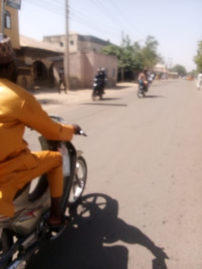 The scene depicts a daylight street view in the city of Gama, Nigeria. In the immediate foreground, a person is seated on a light grey/silver motorcycle, facing away from the viewer. This individual wears a mustard-yellow long-sleeved tunic, matching trousers, and a patterned cap. Their left hand is visible on the motorcycle's handlebar. A prominent, elongated shadow of the person and motorcycle is cast onto the asphalt road surface, indicating strong overhead sunlight.
Further down the road, two additional individuals are visible on motorcycles, moving in the same direction as the foreground subject. The first distant rider appears to wear dark attire and a helmet, while the second wears lighter clothing. The road itself is paved and appears to be a multi-lane thoroughfare.
The background features low-rise buildings constructed from light-colored materials, some exhibiting arched architectural details. Utility poles with overhead lines are present along the road, and sparse vegetation, including trees, is visible further along the street. The sky is bright and clear, with no clouds discernible. The overall image exhibits a soft focus, particularly towards the background.