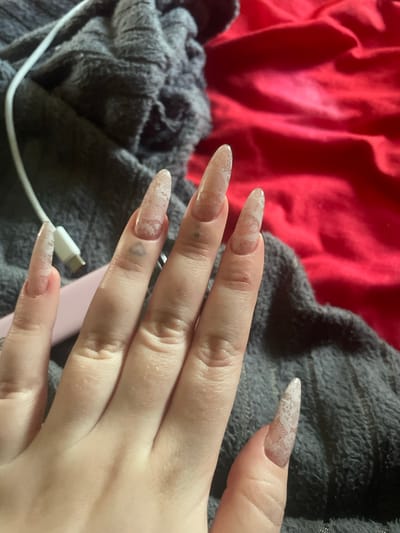A close-up, overhead shot shows a person's left hand with long, pointed, lace-patterned acrylic nails against a backdrop of dark grey and red fabric. A white charging cable with a silver connector is visible on the grey fabric, partially obscuring a pink object. The scene does not provide any information about the location in the city of Линёвка, Russia.