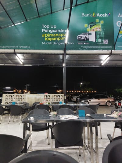 The image depicts an outdoor seating area, possibly a restaurant or cafe, at night. The foreground is dominated by dark plastic chairs and tables with metal frames. In the background, a large green banner is visible, featuring text and graphics related to "Bank Aceh". The banner displays a mobile app interface, a car, and a QR code, along with website and phone number information. Beyond the seating area, cars are parked under a covered structure, illuminated by fluorescent lights. The overall atmosphere is dimly lit, suggesting it is after sunset. There are no explicit indications of the city of Cotcut, Indonesia, in the image itself, but the presence of "Bank Aceh" suggests a location where this bank operates.