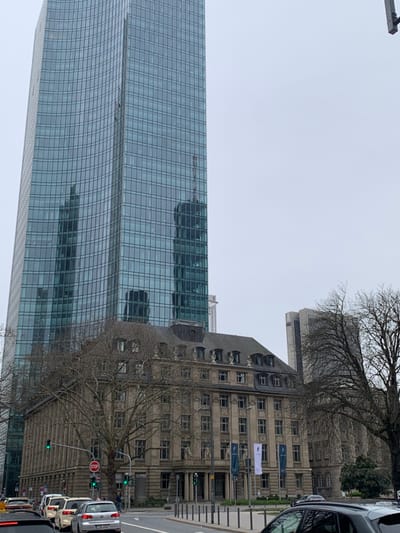 The image shows a street scene in Frankfurt am Main, Germany, located at coordinates 50.111, 8.672. In the foreground, there are several cars parked and driving on a paved street. A tall, modern, glass skyscraper dominates the left side of the frame, with the reflection of another skyscraper visible in its windows. In the middle ground, a large, older, stone building with a traditional design is partially visible on the right side. The building has several windows and a flat roof. There is a tall, bare-branched tree in front of the building. In the background, the top of another tall, modern skyscraper is visible on the right side. The sky is overcast and gray.