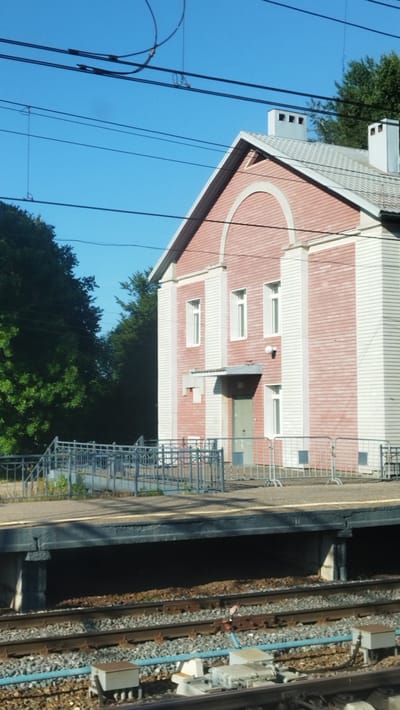 The image shows a train station building and tracks in Vyshny Volochyok, Russia. The building is made of pink and white material and has multiple windows. There are overhead wires and trees in the background. The foreground shows train tracks and a platform.