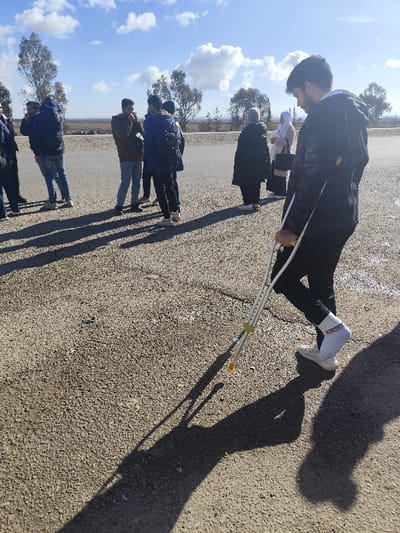 A man with crutches stands in the foreground of a group of people outdoors on a sunny day. He wears a jacket, dark pants, white socks, and sneakers. The ground is a tan gravel surface. Behind him, a group of people stands by a low wall, under a clear blue sky with scattered clouds. Sparse trees dot the horizon. The man's shadow is prominent on the ground in front of him.
