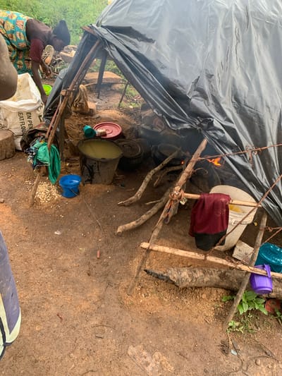 The image shows a makeshift shelter constructed with large black plastic sheeting supported by wooden poles. Inside the shelter, a fire is burning, with several cooking pots placed over it. To the left of the shelter, a woman wearing a colorful patterned skirt and a dark top is tending to something near a large white sack. Various buckets, bowls, and other containers are scattered around the entrance of the shelter. The ground is composed of dirt and small stones. The environment appears to be outdoors, with greenery visible in the background. The scene suggests a basic living or cooking area, possibly in a rural or informal settlement. The location is identified as Buga da Gindi, Nigeria.