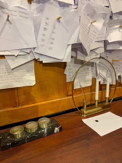 A wooden wall, partially covered with a collection of handwritten notes and messages, is seen to the left of the frame. The wall is mostly a light brown color, with a darker brown strip that runs horizontally along its bottom edge. The notes are of various sizes and shapes, and are arranged in a seemingly random pattern. Several notes are pinned to the wall with small wooden pegs. The notes are written in a variety of languages, including English, Indonesian, and Chinese. Some notes are partially visible, while others are completely obscured by the wall. To the right of the wall is a wooden table, with a brass candle holder containing three white candles. The candles are lit, and their flames are casting a soft glow on the table surface. The candle holder is sitting on a piece of white paper that is partially obscured by the edge of the table. In front of the candle holder are three small, circular candle holders on a black marble base. A dark brown desk, likely wood, is beneath the wall. This is a scene from a place in the city of Kecamatan Borobudur, Indonesia, located at latitude: -7.606, longitude: 110.18.