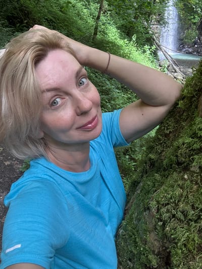 A blonde woman with light blue eyes and fair skin is taking a selfie in a forest setting. She is wearing a bright blue t-shirt and has her left arm raised, with her hand resting on the back of her head. Her right arm is bent and resting on a large tree trunk covered in moss. The woman is smiling at the camera, and her lips are glossy. In the background, a waterfall cascades into a pool of water, surrounded by lush green foliage. There are a few other people visible in the distance near the waterfall. The overall impression is one of a serene outdoor adventure. The image is taken in Plastunka, Russia.
