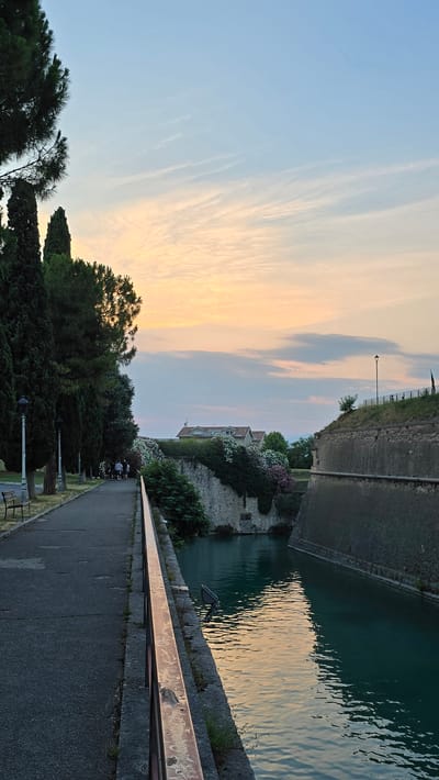 An outdoor, ground-level, vertical shot captures a serene canal in Peschiera del Garda, Italy, during sunset. The sky, a gradient of soft blues and oranges, reflects on the still, teal water. On the left, a tree-lined pedestrian path runs alongside the canal, with a few people strolling and sitting on a bench. A reddish-brown railing separates the path from the water. On the right, the canal is bordered by a stone fortification, its rough texture contrasting with the smooth water. Buildings and lush greenery are visible atop the fortification. The scene exudes tranquility and highlights the historical architecture of the city.