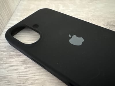 A black iPhone case with a gray Apple logo is placed on a light-colored, weathered wooden surface. The case is positioned at an angle, with the bottom edge closer to the viewer. A circular cutout, likely for a camera lens, is visible on the top left of the case. The wooden surface has a visible grain pattern, suggesting it might be a table or floor. The lighting appears to be natural and somewhat dim, casting subtle shadows. There is no text visible in the image. The scene is static, with the object at rest. The setting is likely indoors, given the lighting and the nature of the object.