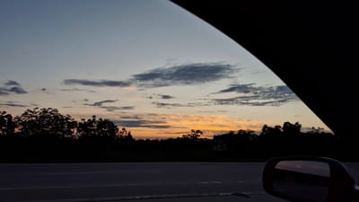This is a photograph taken from inside a vehicle, looking out at a sunset over a rural landscape. The sky is a gradient of pale blue, fading into orange and pink hues near the horizon, with scattered wispy clouds. Silhouetted against the sky are the tops of trees and some buildings, suggesting a village or town. A dark car mirror is visible in the lower right corner of the frame, reflecting the sunset. The foreground shows a road with lane markings, indicating the vehicle is on a paved surface, likely a highway or main road. The overall impression is one of twilight and peaceful transition from day to night. The location is identified as Răzeni, Moldova.