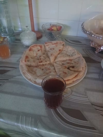 A flat lay photograph shows a breakfast setting on a patterned tablecloth. In the foreground, a small glass cup filled with a dark red liquid, likely tea or juice, is positioned on the right side of the image. To the left and behind the cup, a white plate holds several pieces of flatbread, cut into wedges, with visible green specks, suggesting herbs or vegetables. To the left of the plate, a small glass jar containing an orange liquid, possibly jam or honey, is visible. In the background, various decorative items are arranged: a clear glass jar with a lid, a bowl filled with colorful candies, and a large, ornate bowl with a pearl-like trim. A wooden object, possibly a kitchen roll holder, stands behind the glass jar. The overall scene is set indoors, likely on a kitchen counter or dining table, with a tiled backsplash visible in the upper portion of the image. The lighting is soft and diffused. The image does not contain any information to suggest it is located in Charleville-Mézières, France.