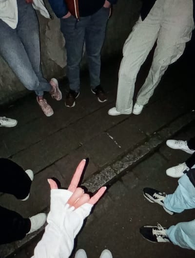 The image depicts a nighttime scene in Aktash, Uzbekistan, showing the lower extremities of at least six individuals gathered outdoors. The ground is a dark paved surface, possibly asphalt or concrete, with a darker, slightly raised strip running horizontally across the mid-ground. A textured, light-colored wall or vertical surface is visible in the background on the left.
From left to right, partially visible individuals include:
1. A person wearing blue jeans and light pink or tan athletic shoes, leaning against the wall.
2. A person wearing dark blue jeans and dark brown athletic shoes, with hands in pockets. A reddish-brown strap is visible near the waist.
3. A person wearing light beige or khaki pants and white athletic shoes.
4. Two individuals (partially visible) wearing light blue jeans and black and white athletic shoes.
5. A person wearing black pants and white athletic shoes.
In the foreground, a left hand is prominently displayed, partially covered by a white long-sleeved garment or glove. The hand is positioned performing the "rock and roll" gesture, with the index and pinky fingers extended upwards, the middle and ring fingers curled, and the thumb extended. The fingernails are long, pointed, and painted a light color. The scene is dimly lit by an unseen artificial light source, casting soft shadows.