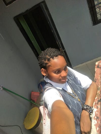 The image captures a female individual, with a dark skin tone, positioned in what appears to be a semi-outdoor or utility area in Cotonou, Benin. The subject, viewed from a slightly low angle, extends her right arm towards the camera, suggesting a self-portrait. She looks subtly to her right. Her hair is styled in short, black cornrows. She is dressed in a white short-sleeved collared shirt beneath a dark blue argyle-patterned sweater vest. A long, thin gold-colored chain with a pendant hangs around her neck. Her left ear displays a small, dark stud earring, and her left wrist is adorned with multiple black beaded bracelets. Direct sunlight illuminates the scene from the upper right, creating strong highlights and shadows. The background on the left consists of a light gray wall with a green pole secured by a white clip. Below the pole, a yellow circular object, a red dustpan, and a broom are observed. In the upper background, two dark, rectangular openings are visible. The far right side of the frame shows a vertical section of a weathered, dark reddish-brown structure with peeling paint. The precise location within Cotonou cannot be ascertained from the visual evidence.