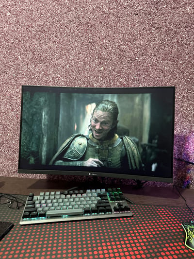 A monitor displays a scene with a man in armor, looking up with a sinister grin. The monitor is black-framed and sits on a surface covered by a textured purple carpet. Adjacent to the monitor on a desk is a gray mechanical keyboard with backlit keys, positioned next to a red and black patterned mousepad. A cable is plugged into the keyboard.
