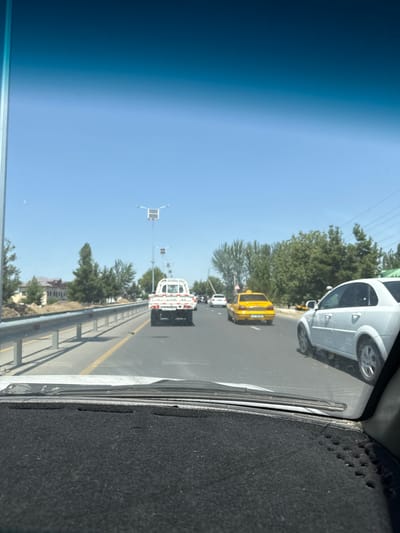 This media file depicts a view from inside a vehicle, looking out through the windshield and side window. The scene is a road with several vehicles, including a white car in the foreground, a yellow taxi, and a white utility truck. Trees and buildings are visible on either side of the road, suggesting an urban or suburban setting. The sky is clear and blue, indicating a sunny day. The orientation of the image is such that the road appears to be running horizontally across the frame. The time of day appears to be daytime, likely late morning or afternoon, given the bright sunlight. The location is identified as Kahramon, Uzbekistan. No specific activity or event is clearly depicted beyond typical road traffic. There is no visible text in the image.