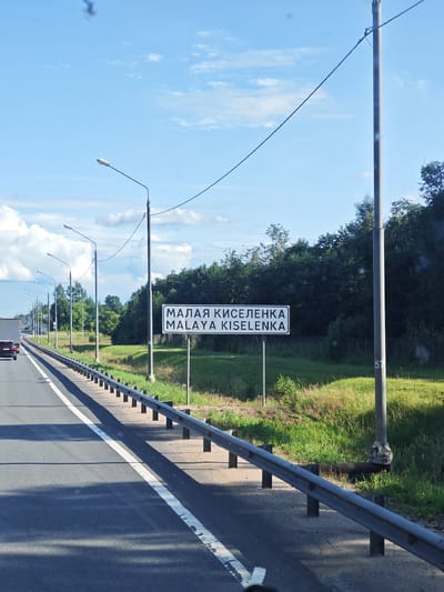 The image depicts a roadside scene in Malaya Kiselenka, Russia. A highway, indicated by a guardrail and lane markings, runs from the foreground towards the background. Several streetlights line the side of the road. A white sign with black text, displaying "МАЛАЯ КИСЕЛЕНКА" and "MALAYA KISELENKA", is prominently visible, identifying the location. A dark-colored car is traveling on the highway in the left lane, moving away from the viewer. The area alongside the highway is characterized by lush green grass and trees, suggesting a rural or suburban environment. The sky is blue with scattered white clouds. A concrete pole with some marking "57" is visible near the sign.