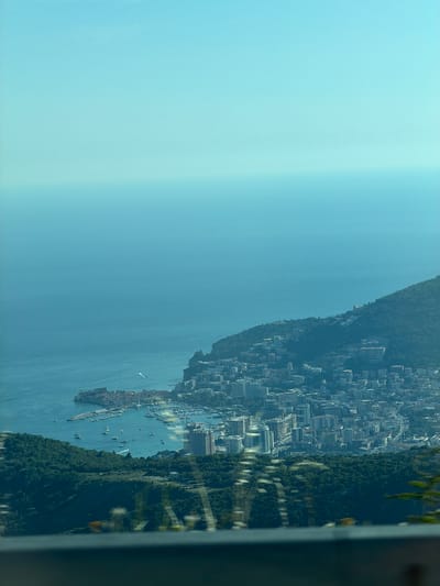 This is a vertical, high-angle, outdoor, medium shot capturing a panoramic view of a coastal city nestled against a lush, green mountainside. The city's architecture, characterized by numerous buildings with white facades and reddish roofs, cascades down the slopes towards the tranquil, blue water of a bay. The bay is dotted with various boats, including sailboats and yachts, suggesting recreational activities or harbor operations. Reflections of the sky and some overhead foliage are visible on the water's surface.
The scene depicts a bright, sunny day with clear skies, likely in the late morning or early afternoon, given the quality of light. The time of year appears to be warm, judging by the vibrant greenery. The location is identified as Ivanovići, Montenegro. The overall atmosphere is serene and picturesque, showcasing a harmonious blend of urban development and natural beauty. The perspective from a higher vantage point emphasizes the scale of the city and its integration with the surrounding landscape. No people are clearly discernible in the frame, and there is no visible text.