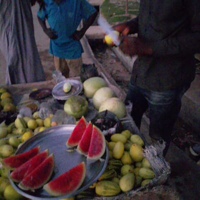 In Wuro Allahamdu, Nigeria, a street vendor displays a variety of fruits. Several slices of watermelon, with their vibrant red flesh and white rind, are arranged on a metal platter. Beside the platter, a pile of small, light-green gourds or cucumbers is visible. In the background, a person wearing a blue garment stands near a stall, and another individual in darker clothing is positioned to the right. A yellow object, possibly a fruit, is held by the person in blue. The scene appears to be outdoors, with a worn, outdoor market setting suggested by the ground surface and scattered debris.