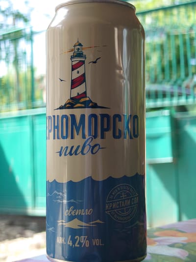 A horizontal close-up shot captures a can of "PHOМОРСКО пиво" (Pomorie Beer), likely photographed outdoors in bright daylight, possibly in Sofronievo, Bulgaria. The can features a design divided into a light cream top section and a dark blue bottom section, separated by white wavy lines suggesting sea waves. A prominent illustration on the cream section depicts a red and white striped lighthouse with a golden top, emitting light rays, accompanied by two black seagulls in flight.
The visible text on the can, in Cyrillic, translates to "PHOМОРСКО пиво" (Pomorie Beer) and "светло" (light/lager), indicating the type of beer. Below the waves, the alcohol content is clearly stated as "АЛК. 4,2% VOL." (ALK. 4.2% VOL.). A distinctive blue emblem on the can includes further text: "ЗА МЕН И ПЛЪТЕН ВКУС" (For me and full taste), and "КРИСТАЛИ СОЛ" (Crystal Salt) with "ОТ ЧЕРНОМОРСКИ СОЛНИЦИ" (From Black Sea Salt Pans), highlighting an ingredient from Bulgaria's Black Sea coast.
The background is softly blurred, showing varying shades of green, suggestive of foliage or a fence, and hints of a colorful, patterned surface, possibly a tablecloth, in the upper right. The overall scene conveys a relaxed, outdoor setting, implying a beverage ready for consumption.