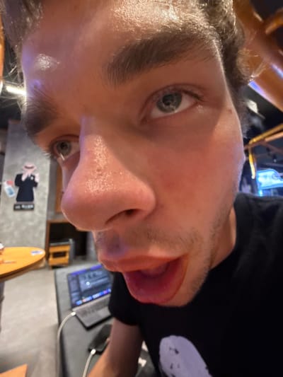 The image is a close-up, rotated portrait of a young adult male, taken indoors in the Minhang District, China. The man's face occupies the majority of the frame, positioned vertically, with his head tilted to the right. He has light-colored eyes, dark eyebrows, and visible stubble on his face. His expression is playful, as he is sticking out his tongue. He wears a dark (likely black) t-shirt.

The background, largely out of focus, reveals an indoor setting, likely a casual workspace or cafe. To the man's right, a laptop computer with a vibrant blue-backlit keyboard is visible on a desk or table. The laptop screen displays vertical bars, possibly representing audio waveforms, code, or a graphic interface. A white cable extends from the laptop. Below the desk, a light-colored, elongated wooden object, resembling a decorative surfboard, leans against a wall or surface. Further in the background, a black silhouette of a figure, possibly a person in a suit, is affixed to a light-colored wall or board. Beneath this silhouette are a few small, indistinct colorful objects and some illegible text. The lighting is artificial and somewhat dim, suggesting the image was taken during the evening or night. The overall mood conveyed by the man's expression is lighthearted and silly.