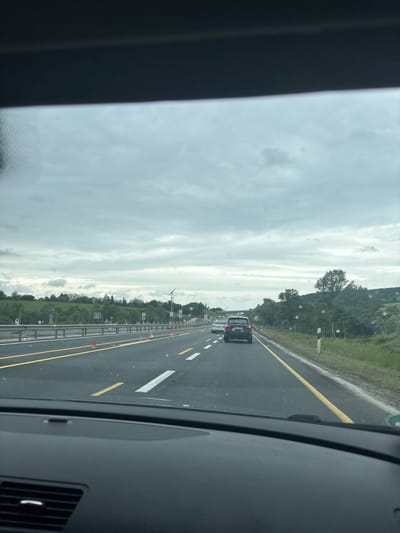 The image, taken from inside a vehicle, shows a view of the M1 highway in Tatabánya, Hungary. The road is divided by solid yellow lines and white lane markers. On the right, a metal barrier runs alongside the road with orange cones visible. Beyond the barrier is a grassy area and trees. Several vehicles, including a dark-colored van, are ahead on the highway. The sky is overcast with visible clouds. The vehicle's dashboard and air vent are visible in the foreground.