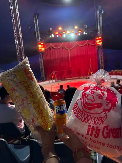 The image shows two bags of popcorn and a can of Fanta being held up, likely in a circus or entertainment venue. The background features a red curtain, stage lights, and a blue tent-like ceiling, suggesting an indoor performance. The popcorn bags have colorful designs; one prominently displays "A HEAP of POPCORN" with cartoon characters, while the other, a clear plastic bag, has a red design of a grinning face and the text "it's Great". The Fanta can is orange and shows the brand name. The overall scene suggests a casual outing, possibly a family event, with people enjoying snacks. The lighting appears to be artificial and set for a performance.