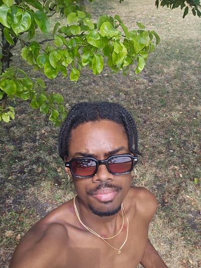 A person with dark skin and braided hair, wearing black sunglasses and a gold chain, is lying on the ground. Their upper body is exposed, and there are visible red marks on their shoulder and neck, resembling lipstick kisses. The person is looking upwards with their mouth slightly open, tongue visible. The background consists of dry grass and fallen leaves, with green foliage from a tree arching into the frame from the right side. The scene appears to be outdoors, possibly in a park or a garden, given the presence of grass and trees. The lighting suggests it might be daytime, possibly overcast as there are no harsh shadows. The overall mood is relaxed and candid.
