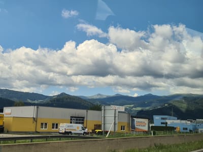 The image shows a commercial building with yellow and grey sections under a partly cloudy blue sky. Mountains covered in trees form the background. A white van with "GO EZY" written on it is parked in front of the building. To the right, there is a large billboard with solar panels and German text. A blue building with "SKF" on it is visible further to the right. The scene is consistent with an industrial or commercial area in Judenburg, Austria, given the mountainous terrain and building architecture.