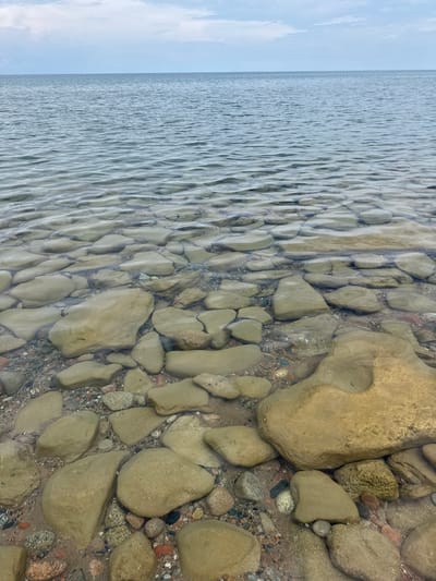 The image depicts a body of water, likely a lake or sea, with a rocky shoreline. The water is clear, revealing numerous rounded stones and pebbles beneath its surface. The stones vary in size, color, and texture, with some appearing smooth and others rough. They are submerged in shallow water, creating a textured underwater landscape. The water itself has gentle ripples, reflecting the sky above, which is overcast with scattered clouds. The horizon line between the water and the sky is distinct. The shoreline is composed of sand and smaller stones, with larger, tan-colored rocks becoming more prominent as they enter the water. The scene is a natural, outdoor environment. There are no people or man-made objects visible in the image. The location is not specified to be in Tong, Kyrgyzstan.