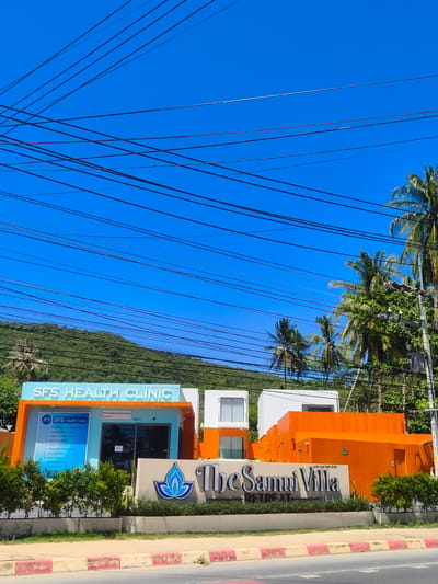 The image shows a sunny exterior view of a modern building complex in Baan Thong Po, Thailand. The main focal point is "SFS Health Clinic," a bright blue and orange building with a sign listing various services. To the right of the clinic, a larger, vibrant orange structure is visible, which appears to be part of "The Samui Villa Retreat," indicated by a large sign. Palm trees and lush green foliage surround the buildings, with a verdant hillside in the background. Overhead, a complex network of power lines crisscrosses the clear blue sky. No people are visible in the scene. The street in front of the complex is paved with red and white curbs.