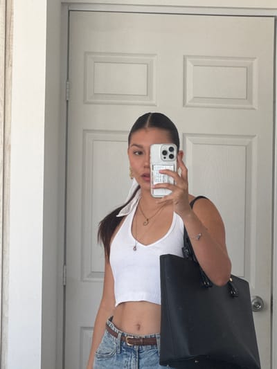 A young woman is taking a selfie in front of a white paneled door. She's wearing a white tank top and blue denim shorts with a brown belt. She's holding a smartphone with a clear case in front of her face. A black tote bag hangs from her shoulder. The location appears to be indoors. The city of San Francisco Ocotlán (Ocotlán), Mexico is not discernible from the image.