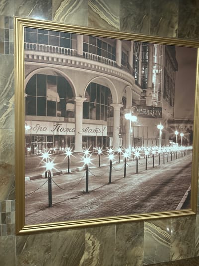 The image shows a framed sepia-toned photograph of a street scene at night, likely a commercial area, possibly a club or restaurant. The photograph is hanging on a wall covered with marbled tiles. The scene depicts an arched building facade with large windows and a prominent sign that reads "CELEBRITY NIGHT CLUB". A row of lampposts with starburst lights lines a cobblestone street, and bollards with chains create a barrier along the sidewalk. There is text in Russian on the building facade that appears to read "Добро Пожаловать", meaning "Welcome". The overall impression is one of a lively night entertainment district.