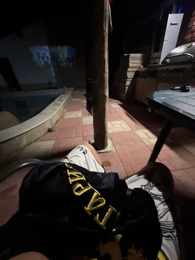 An individual is depicted sitting outdoors at night in Debelt, Bulgaria, with a first-person perspective looking forward and slightly down. The subject's legs are visible in the foreground; the left leg wears white shorts with dark print, and the right leg is partially obscured by a black garment. This garment, likely a jacket or sweatshirt, features prominent gold embroidered letters, possibly "LV" followed by other characters, and a faint graphic of a stylized figure. A patterned white pillow is visible to the right.

The scene includes a patio paved with alternating red and beige square tiles. To the left, a swimming pool with dark water and a light-colored coping is present. A robust, dark wooden pole stands vertically in the midground. To the right, a dark green octagonal table with a dark base holds a small dark object, possibly a mobile phone or remote. Behind the table, a silver-colored barbecue grill with a raised hood is positioned near a stack of wooden panels or planks against a brick wall.

In the background, a light-colored building wall serves as a projection screen for a video display, showing an indistinct scene with multiple figures, suggesting a movie or television show. The environment is dimly lit by the projector and subtle ambient sources, indicating nighttime conditions.