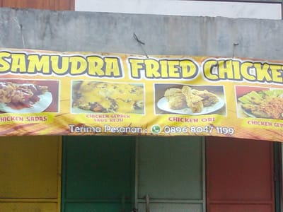The image shows a close-up of a yellow banner advertising "SAMUDRA FRIED CHICKEN". The banner is stretched across the top of a building facade, above a section of wall that appears to be made of concrete or stucco, with some visible stains and wear. The banner features four framed images of fried chicken dishes. The text on the banner includes "CHICKEN SADAS", "CHICKEN GEPREK SAUS KEJU", "CHICKEN ORI", and "CHICKEN GEP". Below the images, the banner displays "Terima Pesanan : C 0896 8047 1199". The bottom portion of the image reveals parts of doors or panels that are red and green, with a yellow panel on the left. The context suggests this is a storefront or food stall in Megamendung, Indonesia, likely a casual dining establishment given the banner's design and the visible doors. There are no people visible in the frame.