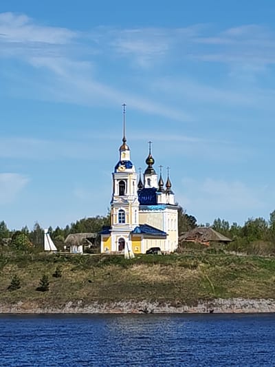 The image shows a yellow and white Russian Orthodox church with blue domes, located in Глебово, Russia. The church sits atop a grassy hill overlooking a body of water. There are trees and other buildings visible around the church, including a small, white, tower-like structure to the left of the church. A black car is parked near the church. The sky is blue with some scattered white clouds.
