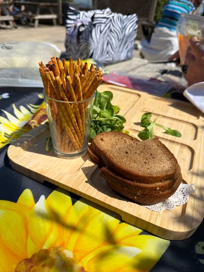 The image displays an outdoor setting in bright daylight, likely at a casual gathering in Monistrol de Montserrat, Spain. The main subjects are food items arranged on a table.
In the foreground, a light-colored wooden serving board with carved compartments holds several slices of dark bread, placed on a white doily. Beside the bread, sprigs of green leafy garnish, possibly lamb's lettuce, are spread. To the left of the board, a clear glass is filled with numerous thin pretzel sticks or breadsticks.
The serving board rests on a black tablecloth featuring a large, vibrant yellow sunflower design. Strong shadows cast by the board and the food indicate bright, direct sunlight, suggesting the time of day is midday or early afternoon.
In the upper left background, a person's hand holds a clear glass or cup. Faint text, appearing to read "Vibert" or "Hibert," is visible on this glass. Further in the background, a chair with a distinctive black and white zebra-stripe pattern is partially visible. The background elements are blurred, suggesting an outdoor environment with other structures. The scene depicts a relaxed meal or snack being enjoyed outdoors.