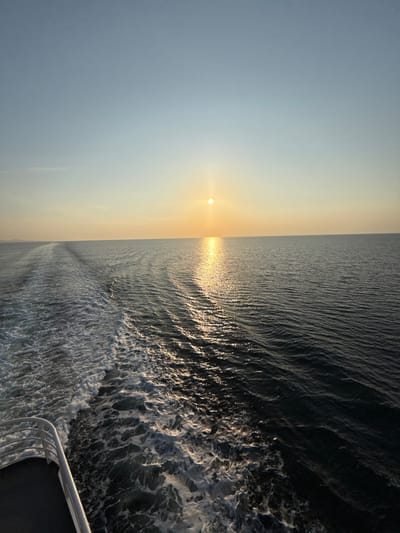 The image displays a wide-angle view of an open body of water at sunset, located in the presumed vicinity of Kato Karioutes, Greece. The horizon line, separating the sky and water, is approximately centered horizontally. The upper portion of the frame features a clear sky, transitioning from a light blue at the top to a warm orange-yellow hue near the horizon. The sun, a bright, circular, golden orb, is positioned low on the horizon, slightly to the right of the frame's center.
A pronounced path of intense golden sunlight reflects directly on the water's surface, extending from the sun towards the foreground, creating shimmering highlights on the waves. The water itself is a deep blue-grey, showing moderate surface ripples. In the lower left quadrant of the image, the white, frothy wake of a moving vessel is clearly visible, extending diagonally from the bottom-left corner towards the distant horizon on the left. This wake signifies the active propulsion of a watercraft. Part of a white metal railing, belonging to the stern or side of the vessel from which the photograph was taken, is visible in the extreme bottom-left corner. No individuals or other discernible objects are present in the frame. A faint, dark landmass is barely discernible on the far left horizon.