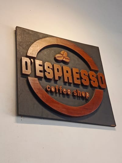 A wooden sign for "D'ESPRESSO coffee shop" is mounted on a white wall. The sign is rectangular and dark gray with a circular cutout in the center. The cutout has a reddish-brown wooden frame. The words "D'ESPRESSO" are carved in raised, reddish-brown wooden letters within the circle, and the words "coffee shop" are carved in smaller, similar letters below. Two coffee beans are depicted in relief above "D'ESPRESSO". The sign appears to be in a coffee shop or similar establishment in Namplohbaro, Indonesia.