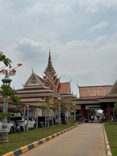 The image shows a building in the Bavet Quarter, Cambodia, featuring a tiered roof with ornate decorations. A road runs alongside the building, bordered by a grassy area and a line of parked cars beneath a shelter structure. People are visible walking along the road in the distance. The sky is overcast.