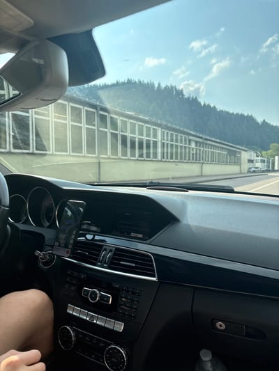 This is a first-person perspective shot from inside a car, looking out through the windshield. The car's dashboard is prominently featured, displaying various controls and a smartphone mounted on a holder. The smartphone screen shows a map application, suggesting navigation is in progress. The time of day appears to be daytime, with bright sunlight filtering through the windshield and casting shadows.
Through the windshield, the scene reveals a highway with a sound barrier running alongside it, separating the road from a forested area on a slope. The sound barrier has a distinctive grid-like structure. Beyond the barrier, dense green trees are visible against a bright blue sky with scattered white clouds. The location context provided places this scene in Gernsbach, Germany. The overall mood seems neutral, consistent with everyday travel. Visible text includes labels on the car's dashboard controls such as "RADIO", "NAVI", "TEL", "AUDIO", "SYSTEM", "MEDIEN", "KLANG", "CLEAR", "ON", "ECO", "A/C", "ZONE", and "AUTO", as well as numbers and letters associated with a keypad. The smartphone screen also displays text related to navigation and potentially song titles.