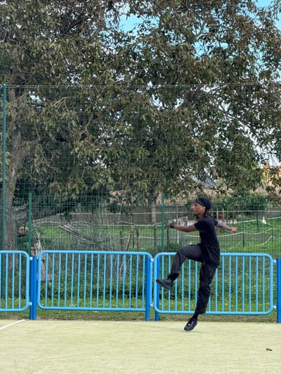 This image captures an outdoor scene on a sports court in Niederhausbergen, France. A male individual is depicted mid-jump, positioned right of center. He has dark skin and braided hair, covered by a black durag. He is wearing a black short-sleeved t-shirt, dark cargo pants, and black athletic shoes. A black watch or wristband is present on his left wrist. His right leg is bent and raised forward, while his left leg is extended downwards, with his foot suspended just above the light green, textured court surface. His arms are bent at the elbows and held outward. In the mid-ground, behind the individual, a bright blue metal barrier with vertical slats runs horizontally across the frame. Behind this barrier, a green chain-link fence encloses a grassy area with sparse vegetation and several large trees featuring dense green foliage, which fill the upper background. A clear, light blue sky is visible through the tree canopy. Faint distant structures or fields are discernible beyond the fence line.